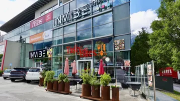 Modernes Geschäftsgebäude mit großer Glasfassade, Café-Eingang und Außensitzbereich vor alpiner Kulisse.