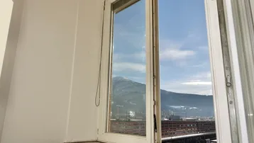 Fensterblick auf eine Berglandschaft mit städtischer Bebauung im Vordergrund unter blauem Himmel.