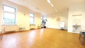 Heller, großer Raum mit Holzfußboden, vielen Fenstern und einer kleinen Spüle, ideal für Therapien oder Seminare.