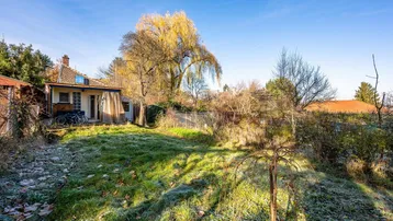 Großer Gartenbereich mit Rasenfläche, Bäumen und einem kleinen Gartenhaus im Hintergrund unter blauem Himmel.