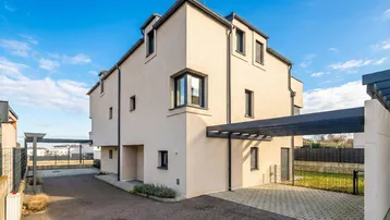 Modernes Reihenhaus mit heller Fassade, Carport und gepflasterter Zufahrt unter blauem Himmel.