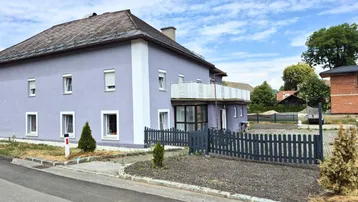 Ansicht des Gebäudes von außen mit lila Fassade und weißem Balkon unter blauem Himmel.