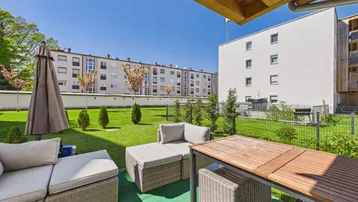Gemütliche Terrasse mit Rattanmöbeln und Blick auf den Gemeinschaftsgarten und umliegende Gebäude.