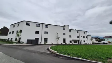 Modernes weißes Gebäude mit schwarzen Fenstern und neu angelegter Außenanlage unter bewölktem Himmel.