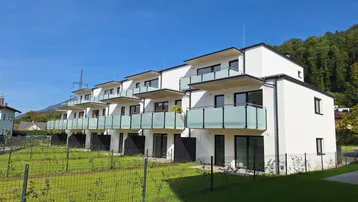 Moderne Reihenhäuser mit weißen Fassaden, Balkonen und kleinen Gärten vor einer grünen Bergkulisse.