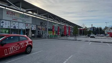 Außenansicht eines Einkaufszentrums mit großem Parkplatz und Geschäften unter bewölktem Himmel.