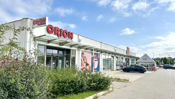 Ansicht der Ladenfront des Orion-Geschäfts mit Parkplätzen und umliegender Vegetation unter bewölktem Himmel.