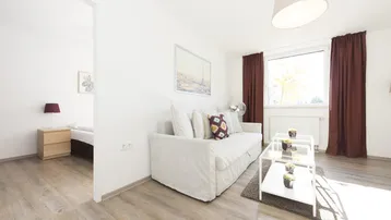 Helles Wohnzimmer mit modernem Sofa, Holzboden und Blick in ein angrenzendes Schlafzimmer.