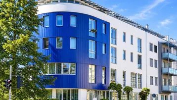 Modernes Gebäude mit auffälliger blauer und weißer Fassade, umgeben von Grünflächen und Bäumen.