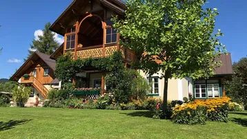 Traditionelles Haus im Landhausstil mit gepflegtem Garten, üppiger Bepflanzung und Holzbalkonen unter blauem Himmel.