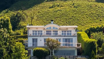 Weiße moderne Villa mit mehreren Balkonen und großen Fenstern, umgeben von grünen Hügeln.