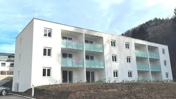 Modernes Mehrfamilienhaus mit weißer Fassade und Balkonen mit Glasbrüstung in grüner Umgebung.