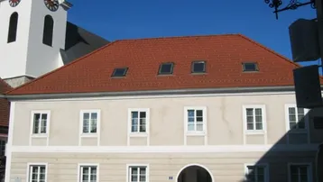 Traditionelles Gebäude mit rotem Ziegeldach und Gaubenfenstern, im Hintergrund eine Kirche.