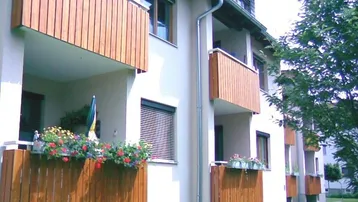 Moderne Gebäudefassade mit mehreren Balkonen, Holzverkleidung und Blumenkästen, umgeben von Grünflächen.