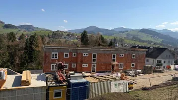 Neubau im Rohbauzustand mit Ziegelmauerwerk und Fensterausschnitten, umgeben von grüner Hügellandschaft.