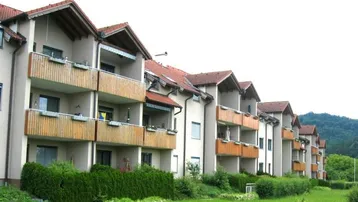 Reihenhausähnliche Wohnanlage mit Balkonen, gepflegtem Rasen und Hecken vor einer grünen Hügellandschaft.
