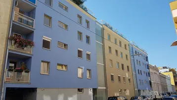 Modernes Wohngebäude mit blauer und beiger Fassade, Balkonen und Dachbegrünung an einer Stadtstraße.