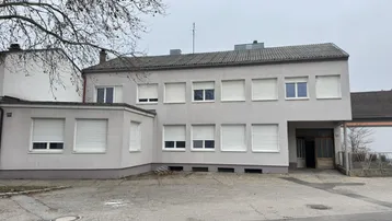 Graues Gebäude mit vielen Fenstern und Rollläden, umgeben von einem kahlen Baum und einem leeren Vorplatz.