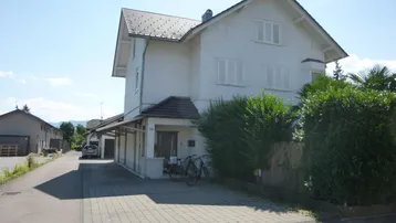 Weißes Wohnhaus mit Satteldach, gepflasterter Einfahrt und grünen Hecken unter blauem Himmel.