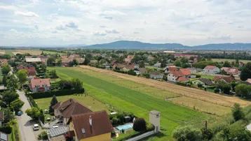 Luftaufnahme einer Wohnsiedlung mit Feldern und Bergen im Hintergrund unter bewölktem Himmel.