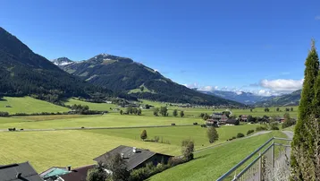 Weitläufiger Blick über grüne Felder und Täler mit majestätischen Bergen im Hintergrund unter blauem Himmel.