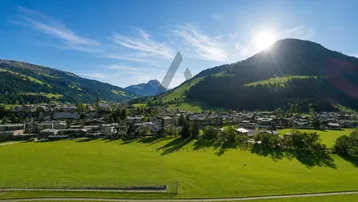 Panoramablick auf ein malerisches Dorf in den Bergen mit weiten grünen Feldern unter strahlendem Sonnenschein.