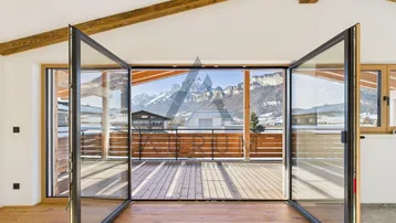 Heller Innenraum mit Holzbalken und großen Glasfronten, die auf eine Terrasse mit Bergblick führen.