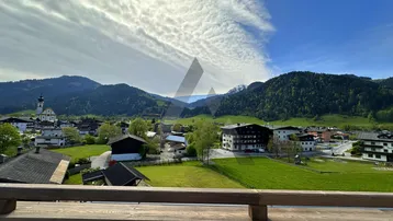 Panoramablick von einem Balkon auf ein malerisches Dorf, umgeben von grünen Hügeln und Bergen.