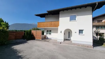Weißes Einfamilienhaus mit Holzbalkon und Bergblick unter blauem Himmel.