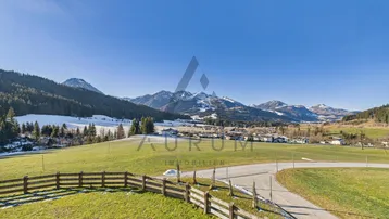 Weitläufige Landschaft mit grünen Wiesen, schneebedeckten Bergen und einem Holzzaun unter blauem Himmel.