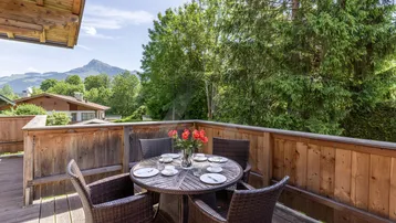 Gemütlicher Balkon mit Holzboden, Esstisch und Stühlen, umgeben von Bäumen und Bergblick.