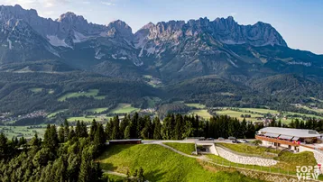 Panoramablick auf ein Gebäudeensemble in den Bergen, umgeben von grünen Hügeln und Wäldern.