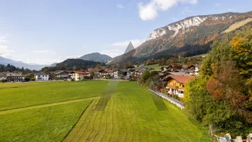Weitläufige grüne Wiesen mit Häusern und Bergen im Hintergrund unter blauem Himmel.