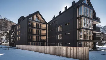 Moderne Architektur mit dunklen Fassaden, großen Glasfronten und Balkonen in verschneiter Berglandschaft.