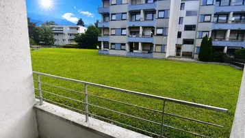 Blick vom Balkon auf eine grüne Rasenfläche und ein Mehrfamilienhaus mit Bergblick.