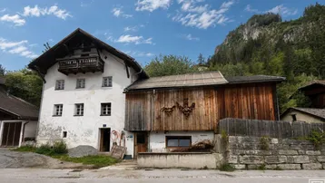 Traditionelles Bauernhaus mit weiß getünchter Fassade und angrenzendem Holzschuppen in ländlicher Umgebung.