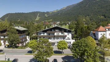 Mehrstöckiges Gebäude mit Balkonen, umgeben von Bäumen und einer beeindruckenden Berglandschaft im Hintergrund.