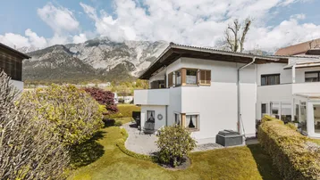 Hausfassade mit gepflasterter Terrasse, grünem Garten und beeindruckendem Bergblick unter blauem Himmel.
