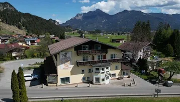 Großes Gebäude im alpinen Stil mit traditioneller Fassadenmalerei, umgeben von grünen Wiesen und Bergen.