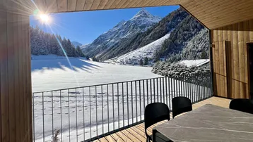 Sonniger Balkon mit Holzboden, Esstisch und atemberaubendem Bergblick auf die verschneite Landschaft.