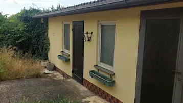 Gelbe Hausfassade mit Fenstern und Türen, umgeben von  überwuchertem Gras und Büschen.