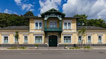 Gelbe Fassade eines historischen Gebäudes mit grünen Akzenten und dekorativem Balkon unter blauem Himmel.