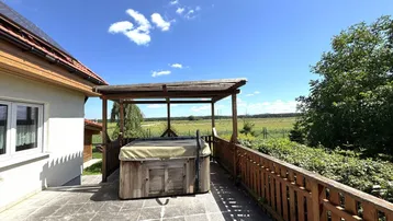 Große gepflasterte Terrasse mit Whirlpool und Holzzaun, umgeben von grüner Landschaft.