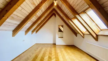 Helles Dachzimmer mit sichtbaren Holzbalken, Holzdecke und Fischgrätparkettboden.