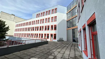 Große gepflasterte Terrasse vor einem modernen Bürogebäude mit roten Fensterrahmen unter blauem Himmel.