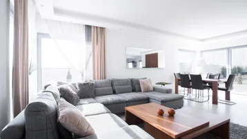 Heller, offener Wohn-Essbereich mit großem Ecksofa, Holztisch und viel Tageslicht durch große Fenster.