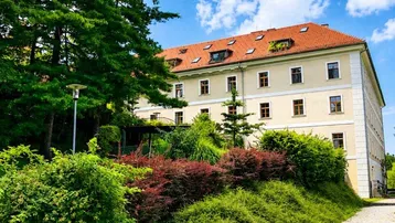 Großes Gebäude mit rotem Ziegeldach, umgeben von üppigem Grün und Bäumen unter blauem Himmel.