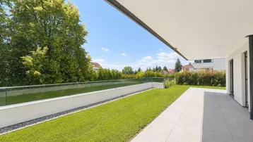 Moderner Gartenbereich mit gepflegtem Rasen, heller Terrasse und Glasgeländer unter blauem Himmel.