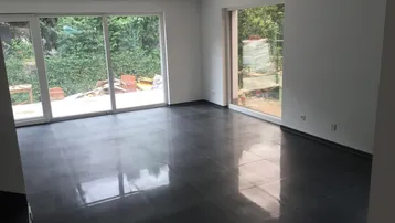 Geräumiges Wohnzimmer mit dunklem Fliesenboden und großen Fensterfronten zum Garten.