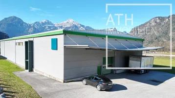 Luftaufnahme eines modernen Gewerbegebäudes mit Laderampe und beeindruckendem Alpenpanorama im Hintergrund.